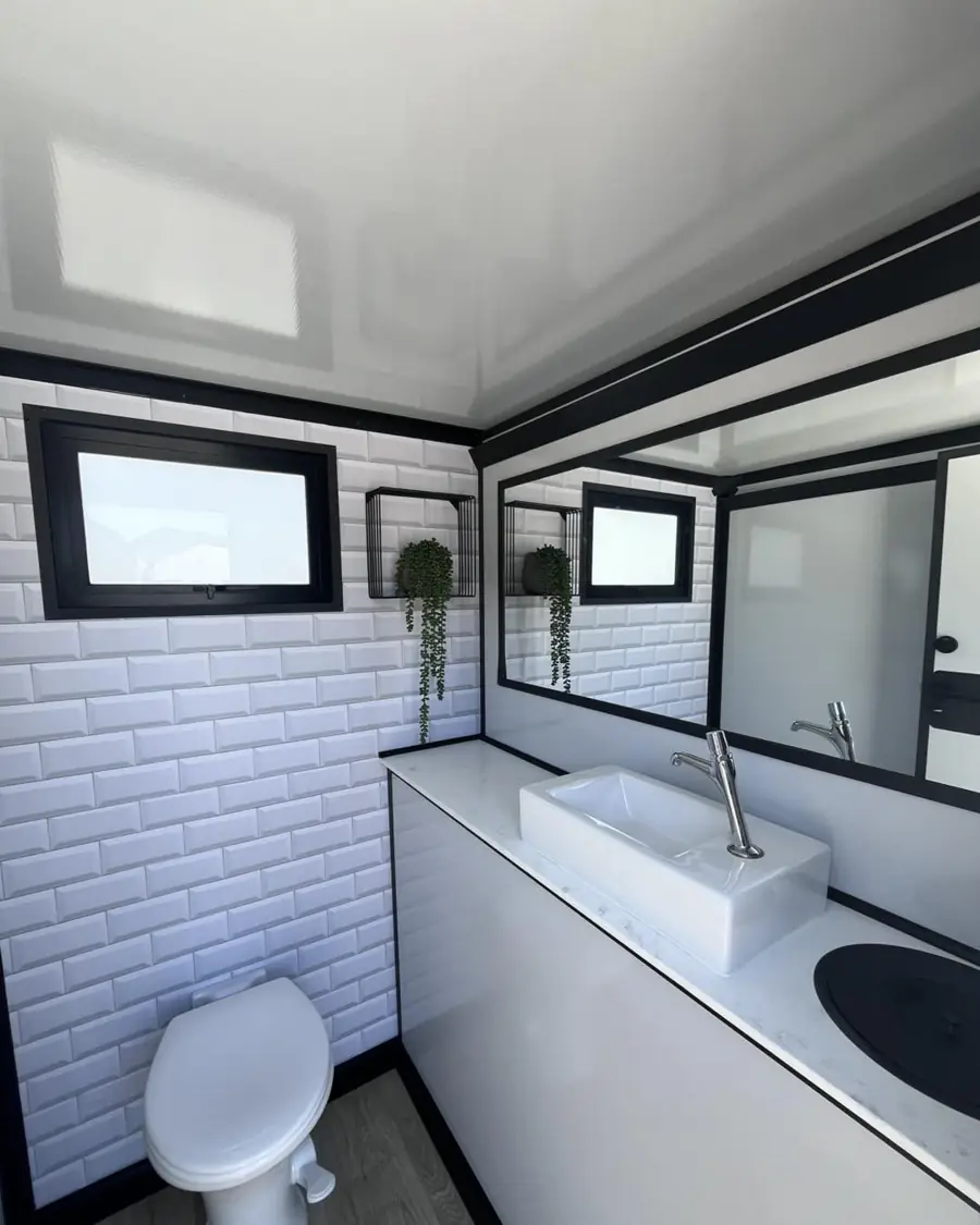 Inside a restroom trailer with a vessel sink, wall mirror, plant shelf and white tile walls